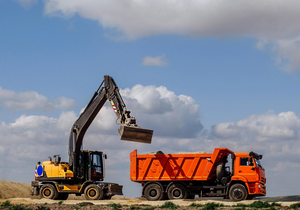 A yellow backhoe loader loads the earth into a truck during the construction of a road against a blue sky with clouds.
